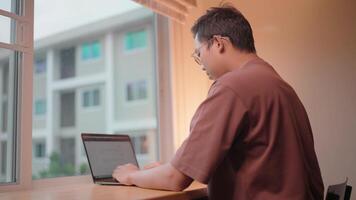 Focus and Determination A man sits at a desk, immersed in work on his laptop, sunlight streaming in through a window behind him. He is surrounded by the comforting familiarity of home. video