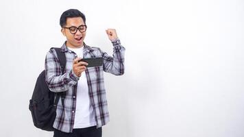 Smiling young Asian male college student with a backpack playing smartphone horizontally isolated on white background photo