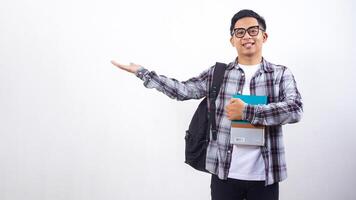 Smiling young Asian male college student with a backpack holding books in front of his chest and presenting empty copy space place isolated on white background photo