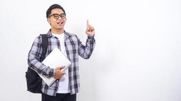 Portrait of smiling young Asian male college student with a backpack and holding laptop netbook while showing gesture of number one isolated on white background photo
