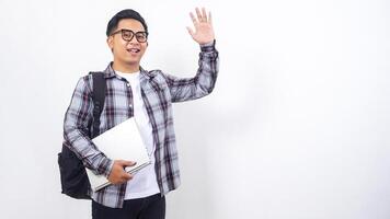 Handsome young Asian male college student with a backpack and holding laptop netbook while showing gesture of greeting isolated on white background photo