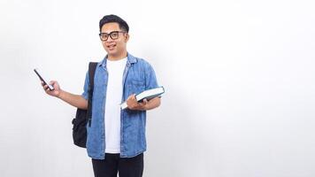 Smiling young Asian male university student in casual outfit with backpack is holding book and smartphone isolated on white background with copy space photo