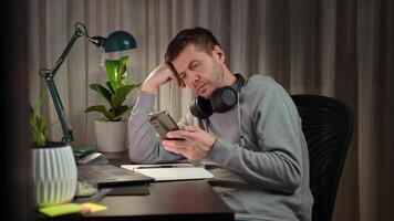 Tired freelancer procrastinating, scrolling phone instead of working. Unproductive man using smartphone at home desk. Concept of procrastination video