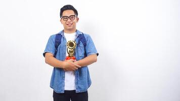 Smiling young Asian male university student in casual outfit with backpack is holding trophy isolated on white background with copy space photo