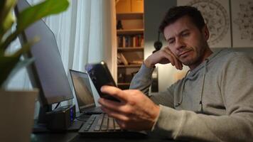 Tired freelancer procrastinating at home office, scrolling phone instead of working. Unproductive man using smartphone at home desk. Concept of procrastination video
