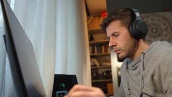 Male freelancer wearing headphones works at computer in home office. Man in gray hoodie working at computer desk with dual monitors and lamp in background. Concept of remote work and productivity video