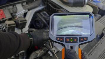 Mechanic inspecting engine cylinders and pistons using borescope camera, live view displayed on handheld screen during car diagnostics under hood in workshop environment video
