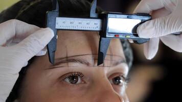 Beautician measuring eyebrows with digital caliper tool on woman's face, symmetrical guideline sketch on forehead for permanent makeup procedure in a professional studio video