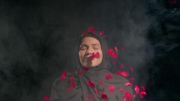 woman in black hijab stands serenely closed eyes as red rose petals gracefully fall around her in soft smoky environment creating captivating and tranquil moment video