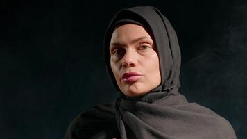 woman dressed in black hijab is captured in indoor setting artistic lighting. She applies cream to face while engaging viewer her expression highlighting her features and attire video