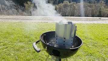 Charcoal chimney starter smoking on a grill outdoors, set against a scenic riverbank, preparing for a barbecue picnic on a grassy riverside under clear daylight video