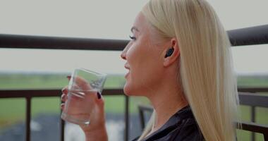 girl stands on a balcony sipping water from a glass. She is engaged in a phone conversation wearing headphones. The backdrop features a scenic view of greenery video