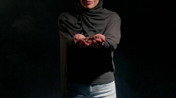 woman in black hijab stands her hands open showcasing small objects in her palms. background is dark highlighting her presence and items she holds video