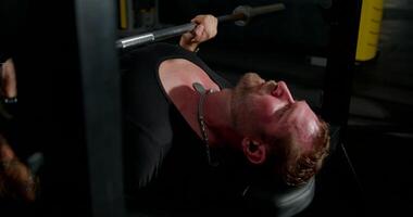 muscular man is focused on lifting weights while performing bench press at a gym. He wears headphones indicating he is listening to music as he works out video