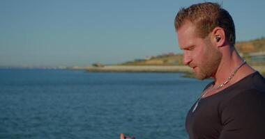 athletic man with a beard is standing by the seashore engaged in a phone call. He wears headphones and is enjoying a sunny day while taking the call near the water's edge video