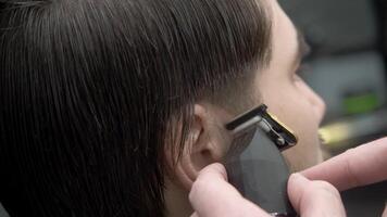 A barber uses a trimmer to give a clear contour and shape to the hair on the temples of a brunette barbershop client. Close up video
