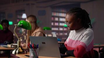 Young scholar focuses on exam preparations in the campus library with colleague, takes notes on laptop from reference books in a peaceful study setting. Girl uses the database materials. Camera A. video