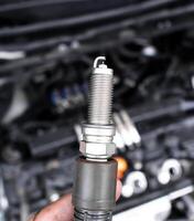 Car Spark Plug, Close up of a single iridium spark plug held in front of a blurred car engine. Automotive ignition system component. photo