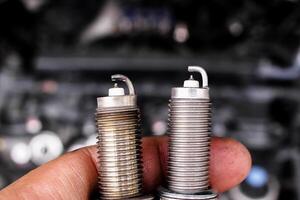 Comparison of used and new spark plugs, A hand holds old and new spark plugs in front of a blurred car engine. Automotive maintenance and ignition parts. photo