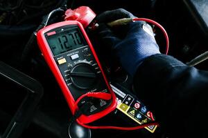 Car Battery Voltage Test, A gloved hand of technician uses a multimeter to measure the voltage of a car battery, displaying a reading of 12.70V DC photo