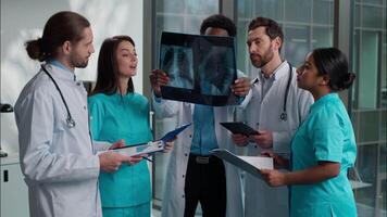 Group of five multiethnic medical students interns men women doctors analyzing patient x-ray result using digital tablet writing clipboard consulting discuss health treatment inside hospital hallway video
