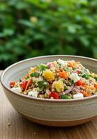 A vibrant bowl of quinoa salad, filled with fresh vegetables and feta cheese, sits on a wooden table. photo