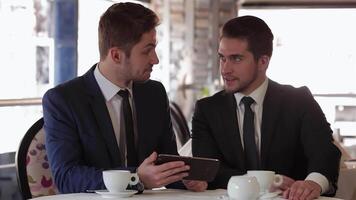 Two men engaged in a focused discussion over a tablet during a business lunch video
