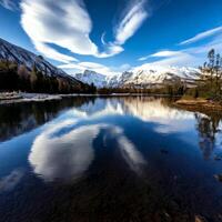 Snow-capped mountains reflecting in calm lake under lenticular clouds photo