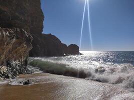 Sunlit ocean landscape waves crashing on a sandy beach, framed by dramatic cliffs under a clear blue sky. Evokes peace and the power of nature. Ideal for travel, escape, or mindfulness themes. photo