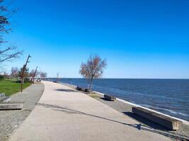 Serene coastal promenade on a sunny day. Empty pathway, benches, and sparse trees offer a tranquil atmosphere. Perfect for travel, tourism, or relaxation themes. photo