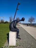 Abstract dark statue of a pensive figure seated on a stone plinth along a sunny promenade. Captures themes of contemplation, solitude, and urban art. photo