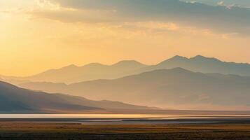 Layered mountain range fading into a hazy, golden sunset or sunrise sky. Still water reflects the light in a vast, tranquil landscape photo