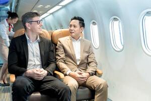 Two businessmen sitting and talking on airplane seats during flight, enjoying business travel experience, with flight attendant working in background cabin aisle photo