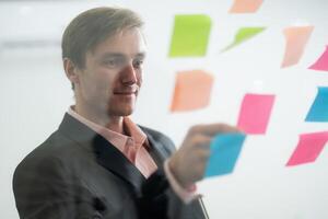 Professional man organizing colorful sticky notes on a glass wall during a brainstorming session at a modern office photo