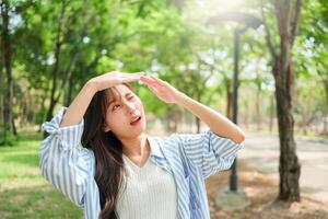 Young Asian woman walking in the park having sunstroke in summer hot weather, Young pretty girl drying sweat using a wipe on a warm summer day in a park photo