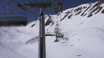 A ski lift moves upward on a snowy mountain slope in the Alps, with enclosed gondolas carrying passengers. Rugged terrain and blue sky are visible. video