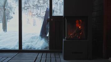 A modern fireplace emits a warm glow inside a cozy room. Snow blankets the ground outside, visible through large glass doors, with flickering flames adding motion. video