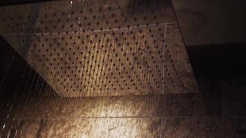 Water cascades from a modern rainfall showerhead against a textured, warmly lit wall, creating a serene and elegant indoor ambiance. video
