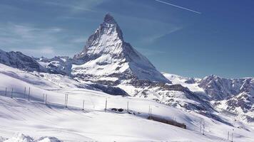 A red Gornergrat train travels along a railway at the base of the Matterhorn in the Swiss Alps, surrounded by snow covered landscapes under a clear blue sky. video