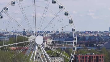 A Ferris wheel rotates slowly in Riga Victory Park, surrounded by greenery. A cityscape with modern and historic buildings and a river is visible. video