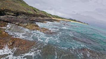 Calm turquoise ocean waves lap against dark volcanic rocks near a sandy beach with lush greenery and a steep hillside in Hawaii. video