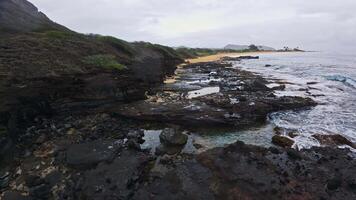 A tranquil beach with orange sand, a rocky mountain, and gentle waves. A person sits on the sand, with palm trees and parked vehicles in the distance. video
