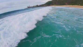 Aerial view of turquoise ocean waters with gentle waves, a sandy beach, lush green hills, and a white tower in a tropical Hawaii setting. video