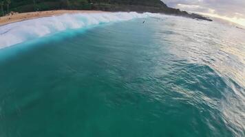 Turquoise ocean waves move gently toward a sandy shore with swimmers visible. A green hillside and coastal community form the backdrop in Hawaii. video