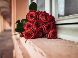 Bouquet of red roses sits on a ledge next to a window photo
