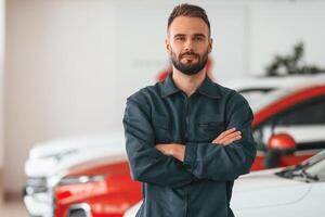 With arms crossed, standing. Car repairman is in the garage with automobile photo