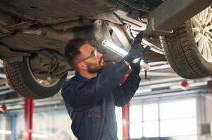 In blue colored uniform. Car repairman ,,is in the garage with automobile photo