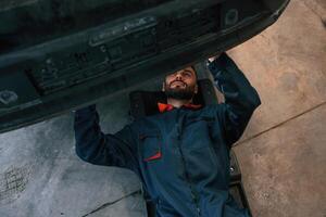 Lying down on the ground. Car repairman is in the garage with automobile photo