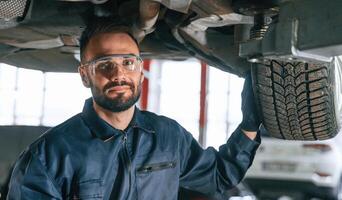 In protective eyewear, under vehicle.,, Car repairman is in the garage with automobile photo