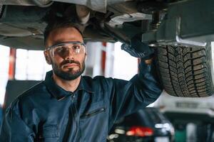 In protective eyewear, under vehicle,. Car repairman is in the garage with automobile photo